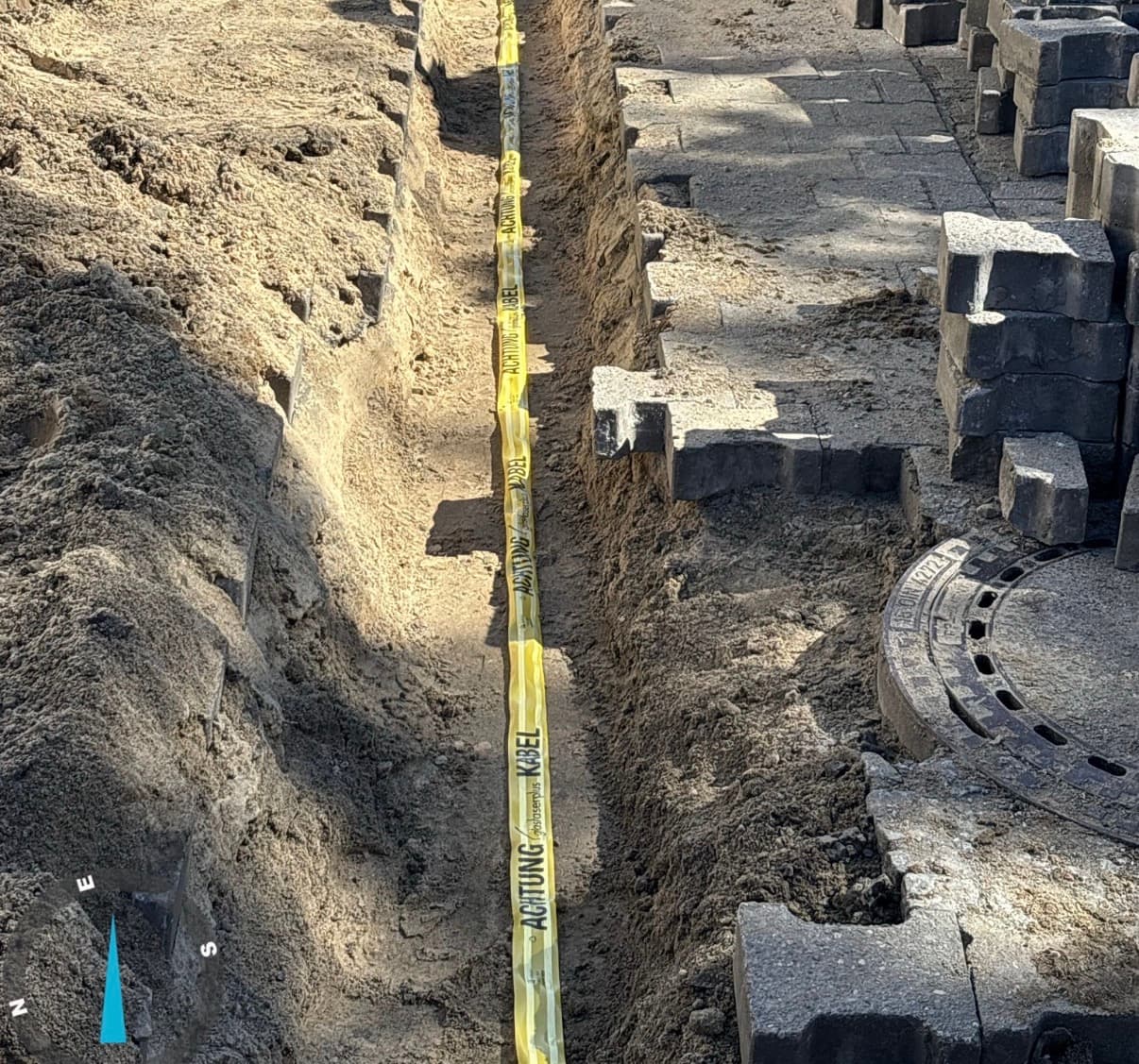 Kabeltrasse mit Warnband im Graben neben Pflasterfläche